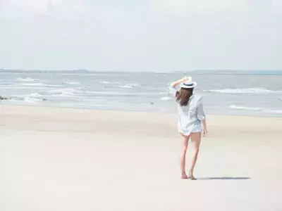A woman holding her hat on her head while standing on the beach.