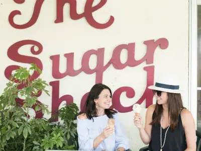 Two women sitting outdoors enjoying an ice cream cone with the sign “The Sugar Shack” in the background.